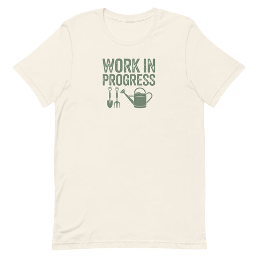 “Work in Progress” gardening graphic with shovel, pitchfork, and watering can printed in sage green on a soft off-white cream colored crewneck t-shirt.