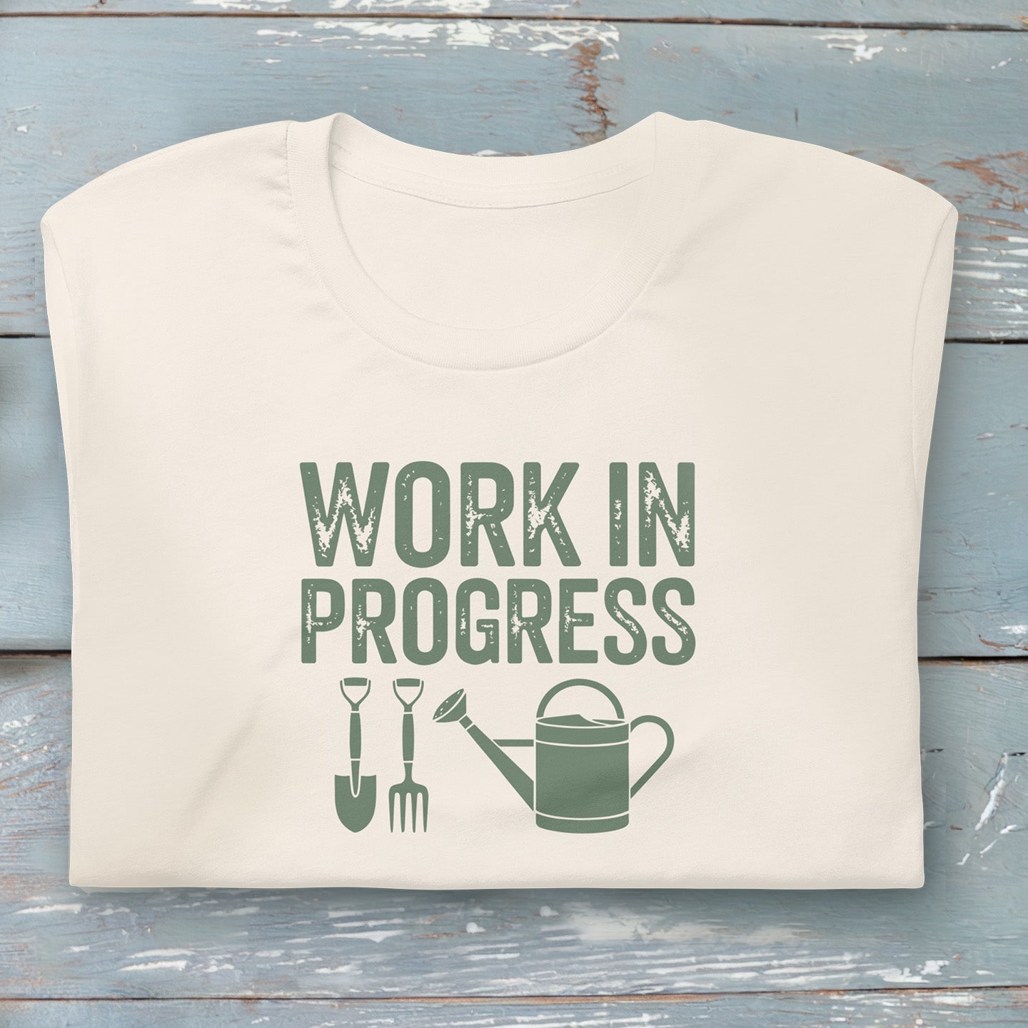 “Work in Progress” gardening graphic with shovel, pitchfork, and watering can printed in sage green on a soft off white cream colored crewneck t-shirt.