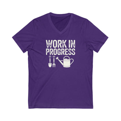 Rich purple colored vneck t-shirt with white words that say 'Work in Progress' in distressed style text. Below the words are graphic images of gardening tools including a shovel, pitchfork and watering can.