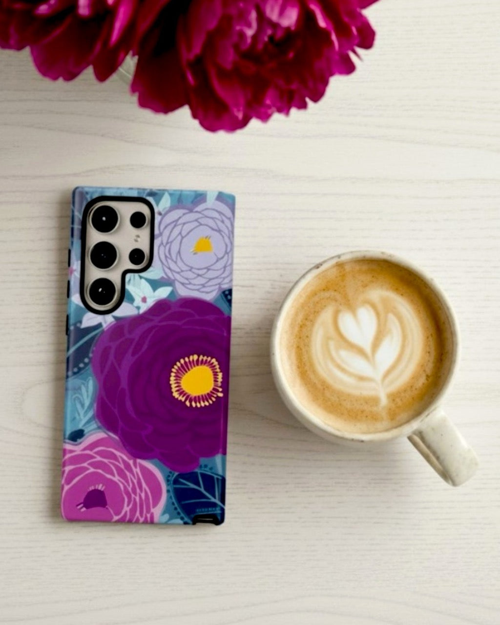 Samsung Galaxy S26 Ultra phone case with Bloomerie violet purple floral design next to a cup of coffee and a pink peony flower on a light surface. Original artwork by designer Kate Spain.