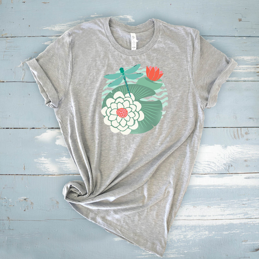 Illustrated dragonfly hovering over a green lily pad with pink and white water lily flowers on a calm pond, printed on a soft athletic gray colored crewneck t-shirt.