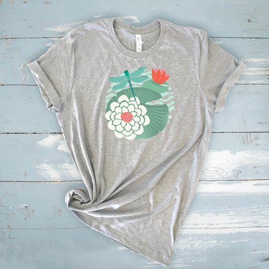 Illustrated dragonfly hovering over a green lily pad with pink and white water lily flowers on a calm pond, printed on a soft unisex t-shirt.