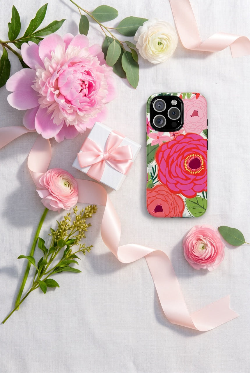 A floral smartphone case with large pink and red flowers and green leaves, shown on a phone in a styled flat lay with pink roses, greenery, a small pink ribbon gift box, and a white fabric background.