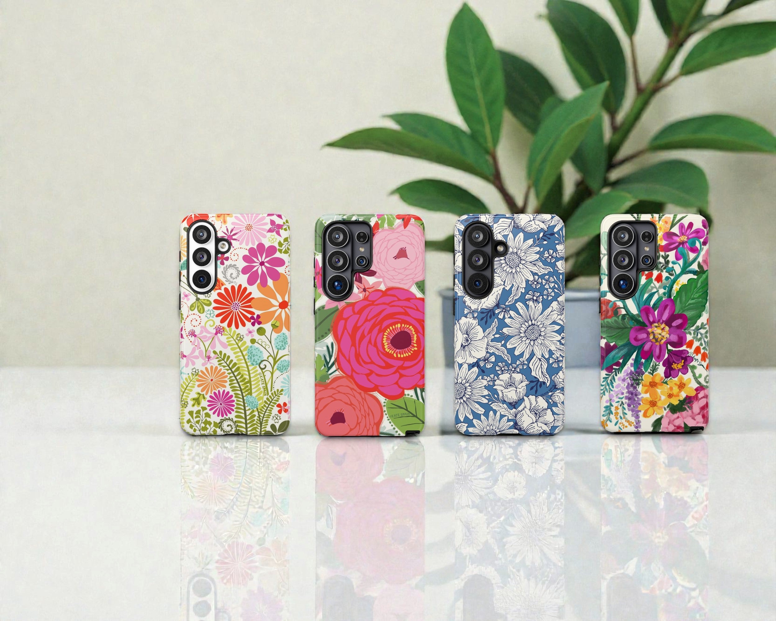Four floral phone cases on a reflective surface with a plant in the background