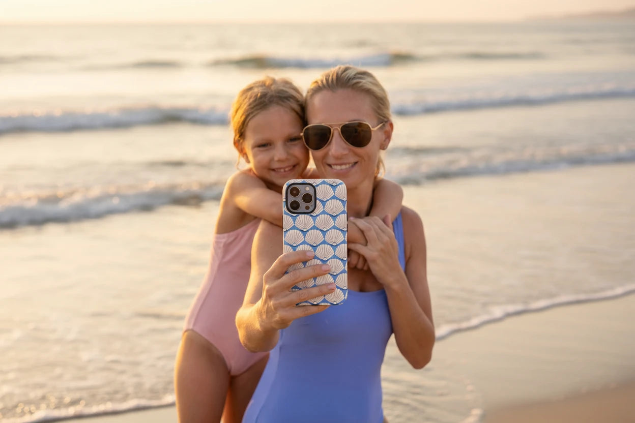 Woman and child taking a selfie on a beach with an iPhone 16 Pro Max phone case featuring a shell pattern. This is the Seashells iPhone case from floriori.