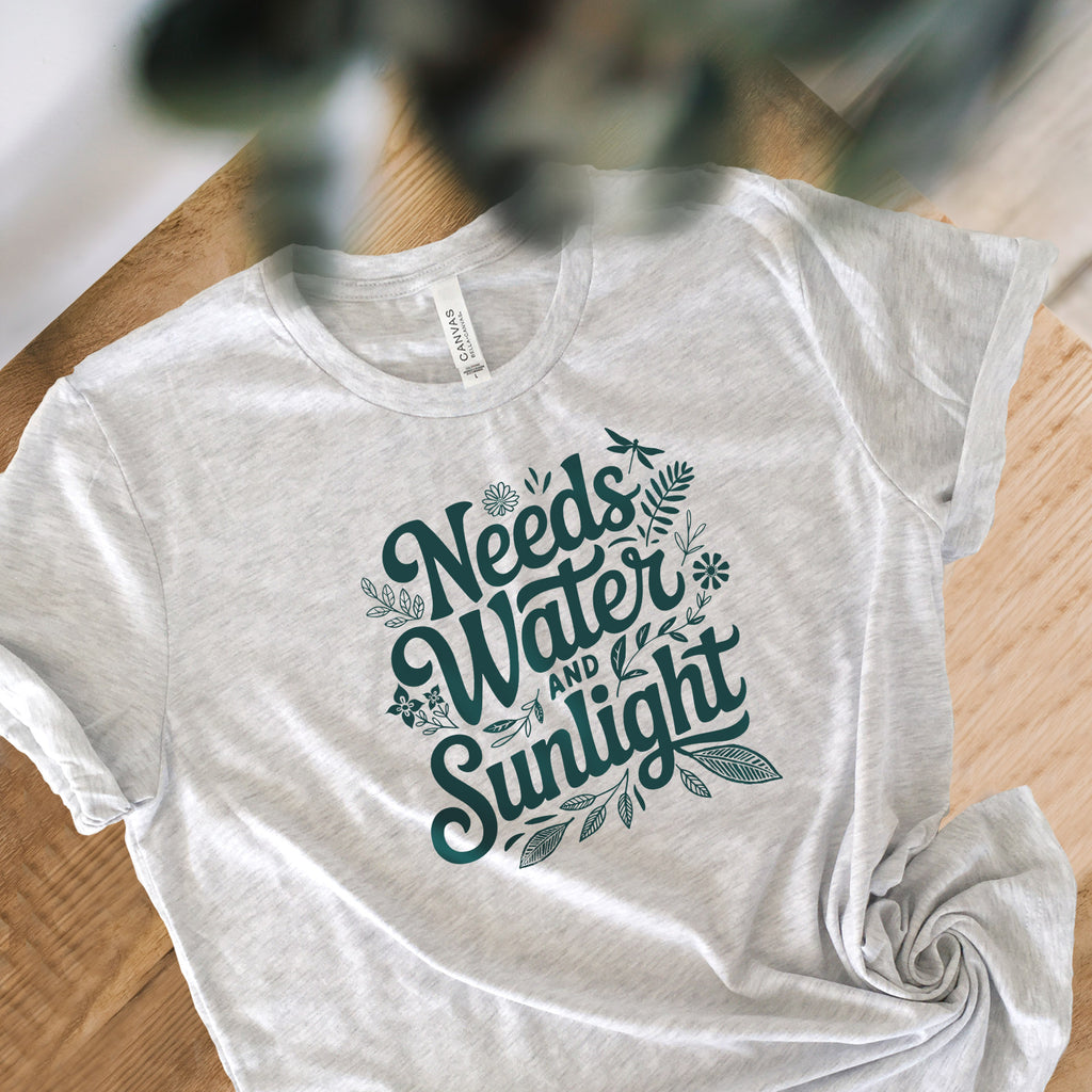 Pale Athletic gray colored crewneck t-shirt with dark green words that say 'Needs Water and Sunlight' in script style text with dragonfly, flower and leaf illustrations around the words.