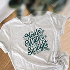Pale Athletic gray colored crewneck t-shirt with dark green words that say 'Needs Water and Sunlight' in script style text with dragonfly, flower and leaf illustrations around the words.