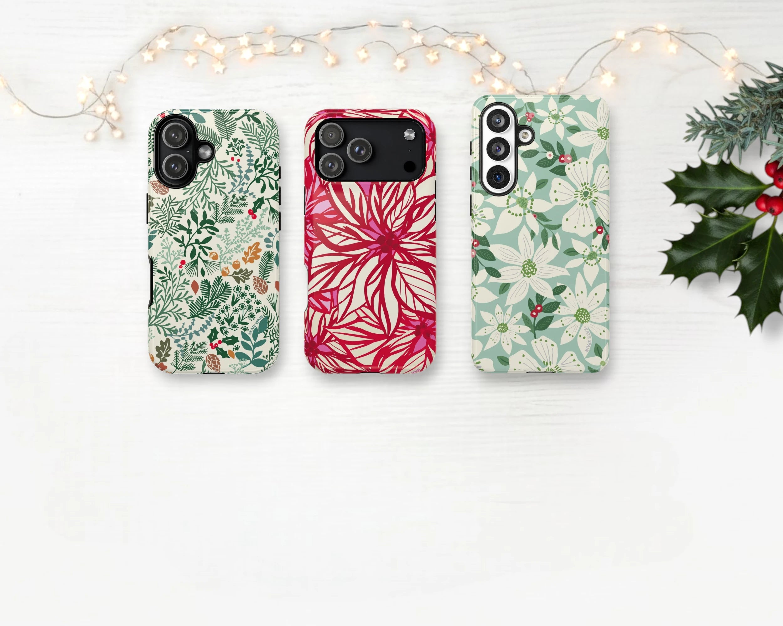 Three festive holiday iphone and samsung galaxy phone cases on a white surface with decorative lights and holly leaves.