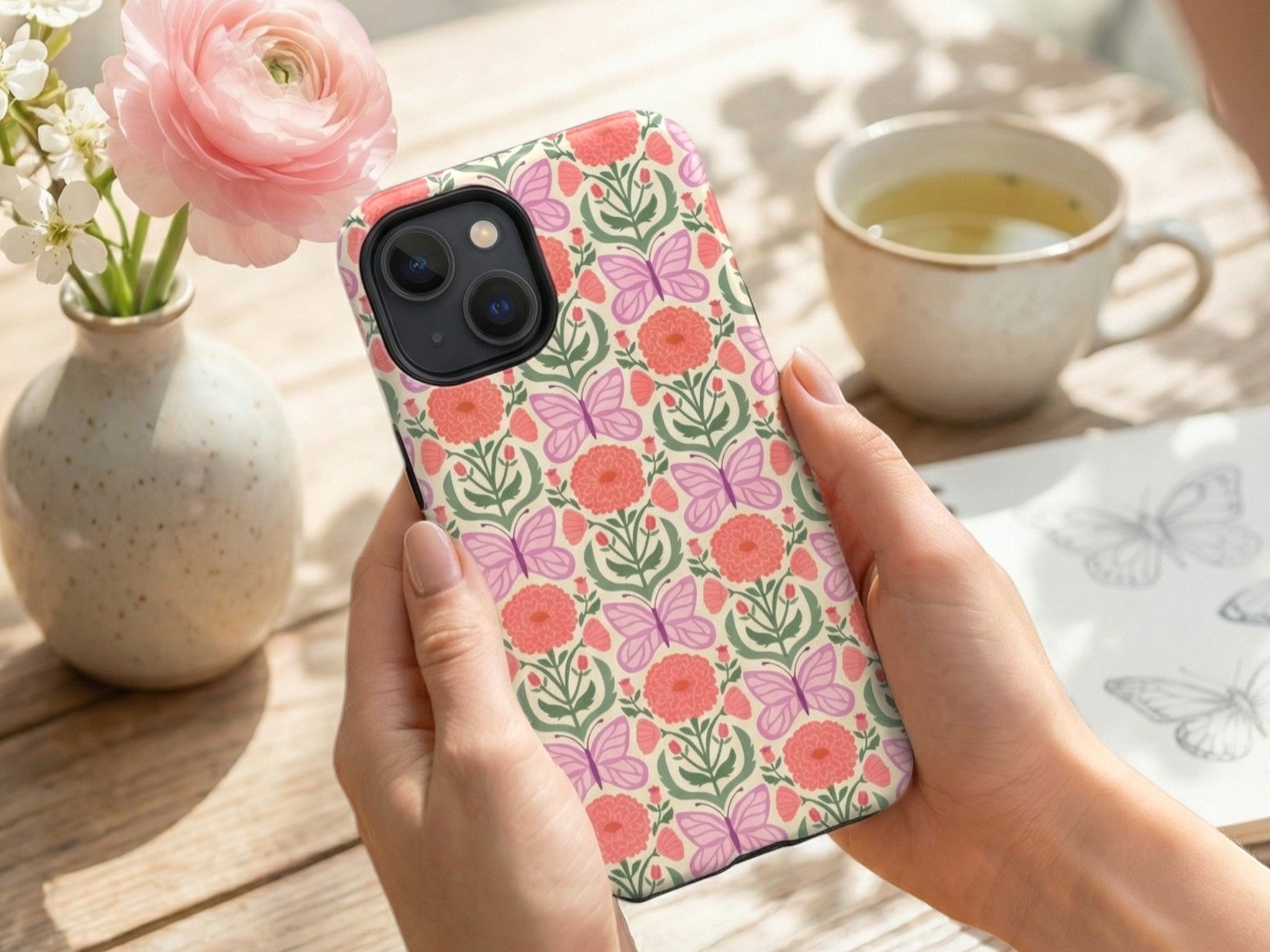 Hand holding a phone case with floral design on a wooden table with flowers and tea.