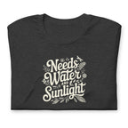 Dark charcoal gray heather colored crewneck t-shirt with white words that say 'Needs Water and Sunlight' in script style text with dragonfly, flower and leaf illustrations around the words.
