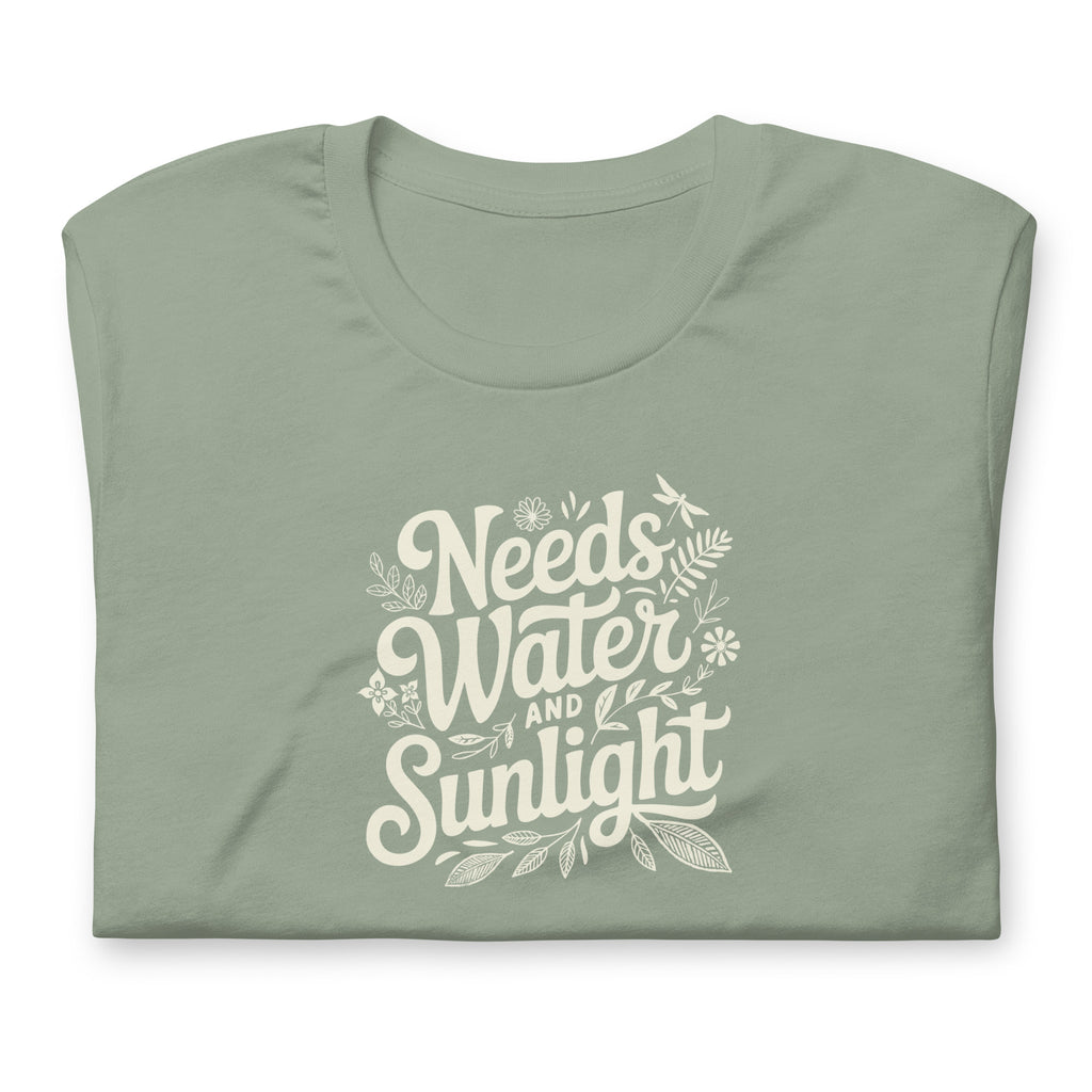 Pale sage green colored crewneck t-shirt with white words that say 'Needs Water and Sunlight' in script style text with dragonfly, flower and leaf illustrations around the words.