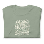 Pale sage green colored crewneck t-shirt with white words that say 'Needs Water and Sunlight' in script style text with dragonfly, flower and leaf illustrations around the words.
