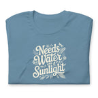 Steel blue colored crewneck t-shirt with white words that say 'Needs Water and Sunlight' in script style text with dragonfly, flower and leaf illustrations around the words.