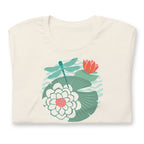 Illustrated dragonfly hovering over a green lily pad with pink and white water lily flowers on a calm pond, printed on a soft off-white cream colored premium quality crewneck t-shirt. Shown folded for scale.
