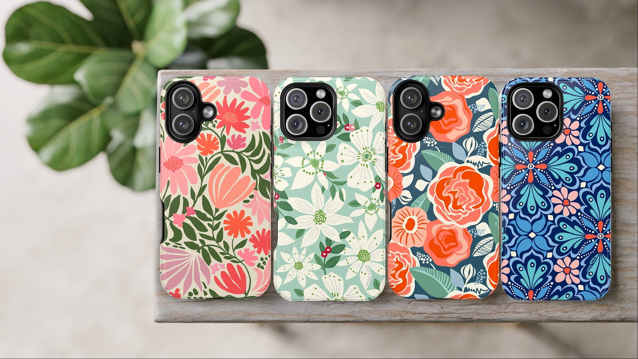 Four floral-patterned phone cases on a wooden surface with a plant in the background.