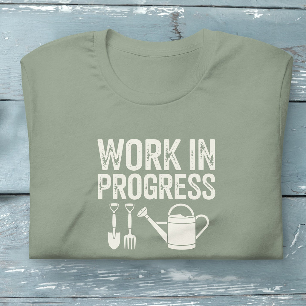 “Work in Progress” gardening graphic with shovel, pitchfork, and watering can printed on a soft sage green crewneck t-shirt.
