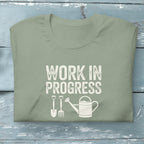 “Work in Progress” gardening graphic with shovel, pitchfork, and watering can printed on a soft sage green crewneck t-shirt.