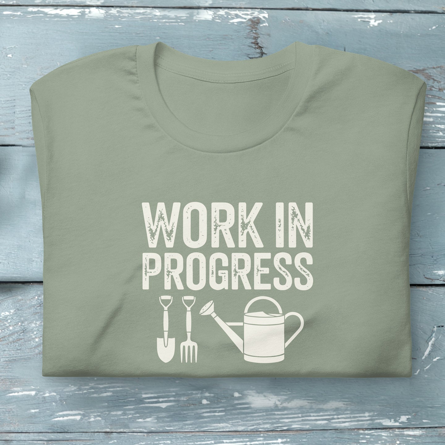 “Work in Progress” gardening graphic with shovel, pitchfork, and watering can printed on a soft sage green crewneck t-shirt.