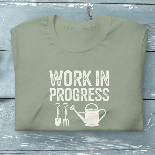 “Work in Progress” gardening graphic with shovel, pitchfork, and watering can printed on a soft sage green crewneck t-shirt.
